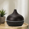 Salt Aroom Suola-Aroma Diffuuseri Aava; tumma, puukuvioitu aromiterapiadiffuusori seisoo vaalealla puupöydällä pienen ruukkukasvin vieressä, taustalla pehmeää luonnonvaloa; diffuusori on mattapintainen ja pyöreä, ei näkyviä painikkeita tai etikettejä pinnalla; ruukkukasvi valkoisessa keraamisessa ruukussa; taustalla epätarkka ikkuna tai seinä; ei ihmisiä kuvassa.