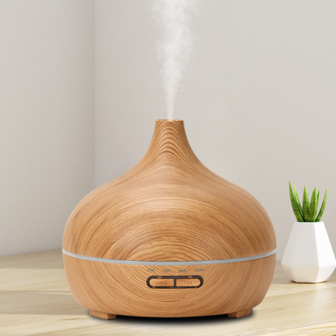 Salt Aroom Suola-Aroma Diffuuseri Aava; puukuvioitu ultraäänidiffuusori päästää höyryä vaalealla pöydällä; diffuusori on pyöreä, mattapintainen, ilman näkyvää merkkiä tai etikettiä; pieni vihreä ruukkukasvi diffuusorin takana, beige seinä taustalla; ei ihmisiä, käsiä tai pakkausta näkyvissä tässä tuotekuvassa.