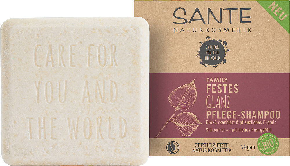 Sante Family Solid Shine Care Palahampoo; kiinteä shampoopala, jonka mattapintaan on kohokuvioitu "Care for you and the world", lepää suorakulmaisen pahvilaatikon vieressä merkinnällä "SANTE Naturkosmetik Family Festes Glanz Pflege-Shampoo"; pakkauksessa lehtikuvituksia sekä vegaani- ja luomusertifiointikuvakkeita; laatikon typografia on lihavoitua groteskia; ei ihmisiä tai toimintaa näkyvissä; tausta yksivärinen ja neutraali.