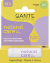 Sante Natural Care Huulivoide; rasia Sante Natural Care -huulivoidetta, merkitty vegaaniseksi ja luomuksi, violetti-keltainen ulkoasu; pakkaus korostaa jojobaöljyä, hyaluronihappoa ja kosteuttavia hyötyjä pehmeille huulille; huulivoidepuikko näkyvissä alaosassa; suorakulmainen rasia mattapinnalla, selkeä lihavoitu tuotenimi groteskikirjasimella keskellä etupaneelia; ei ihmisiä tai taustaelementtejä, kuva otettu suoraan edestä tasaisessa valaistuksessa.