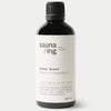 sauna/ing Sauna Scent Spruce & Grape; musta mattapintainen lieriömäinen pullo valkoisella etiketillä; etiketissä mustaa groteskitekstiä: sauna/ing from Finland, Sauna Scent, Spruce & Grapefruit, koordinaatit ja ainesosaluettelo; pullon koko 100 ml; ei ihmisiä tai käsiä; neutraali tausta pehmeässä valaistuksessa studiokuvassa.