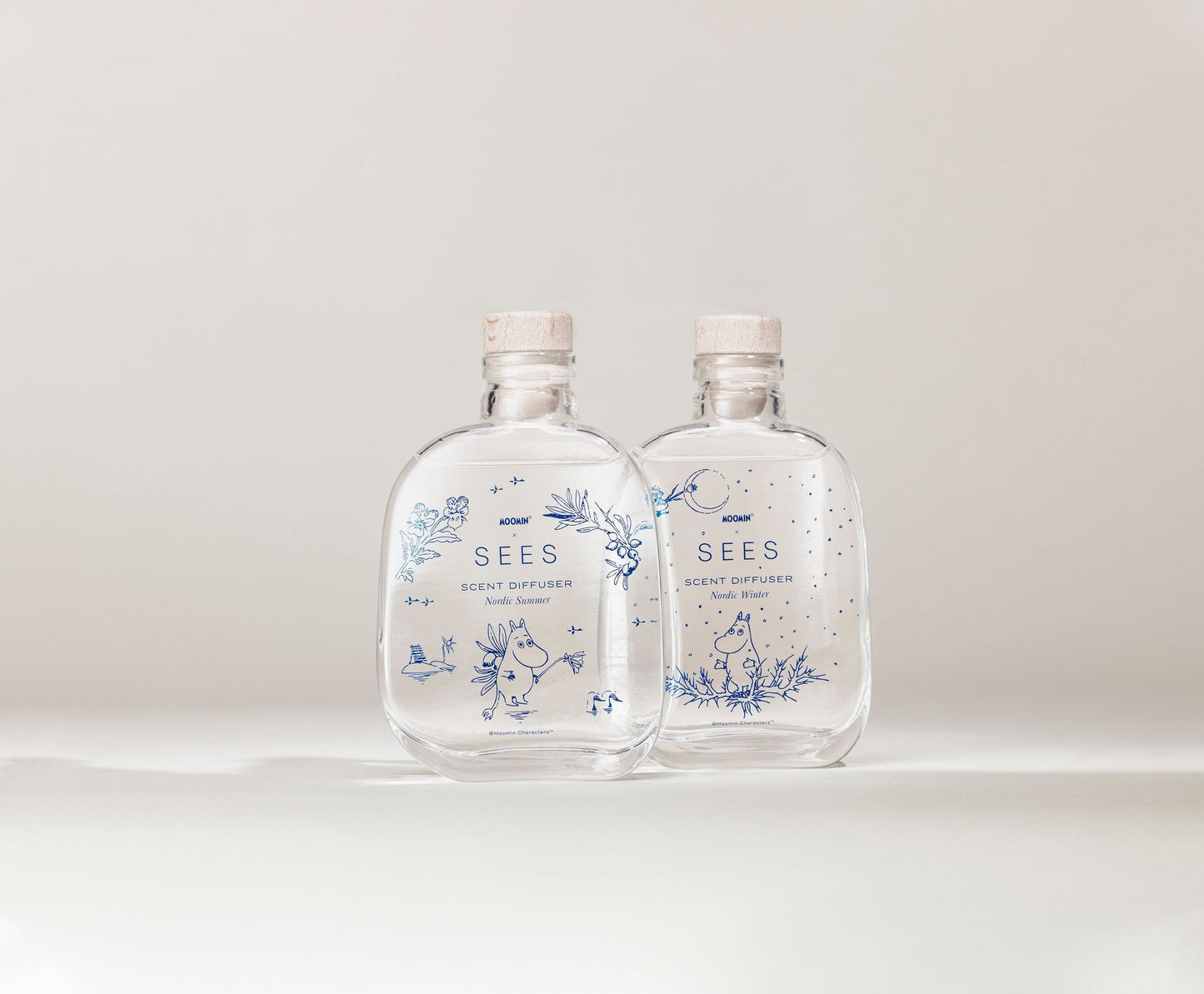 SEES x Moomin Room Diffuser Nordic Winter; kaksi kirkasta lasipulloa korkkitulpin, tekstillä SEES Scent Diffuser; kummassakin pullossa sinisiä kukka- ja pieneläinkuvituksia pinnalla; pullot läpinäkyviä ja lieriömäisiä, mattapaperiset etiketit groteskifontilla; tausta yhtenäinen valkoinen ilman muita esineitä tai tekstuureja; ei ihmisiä tai käsiä, suoraviivainen tuotekuva.