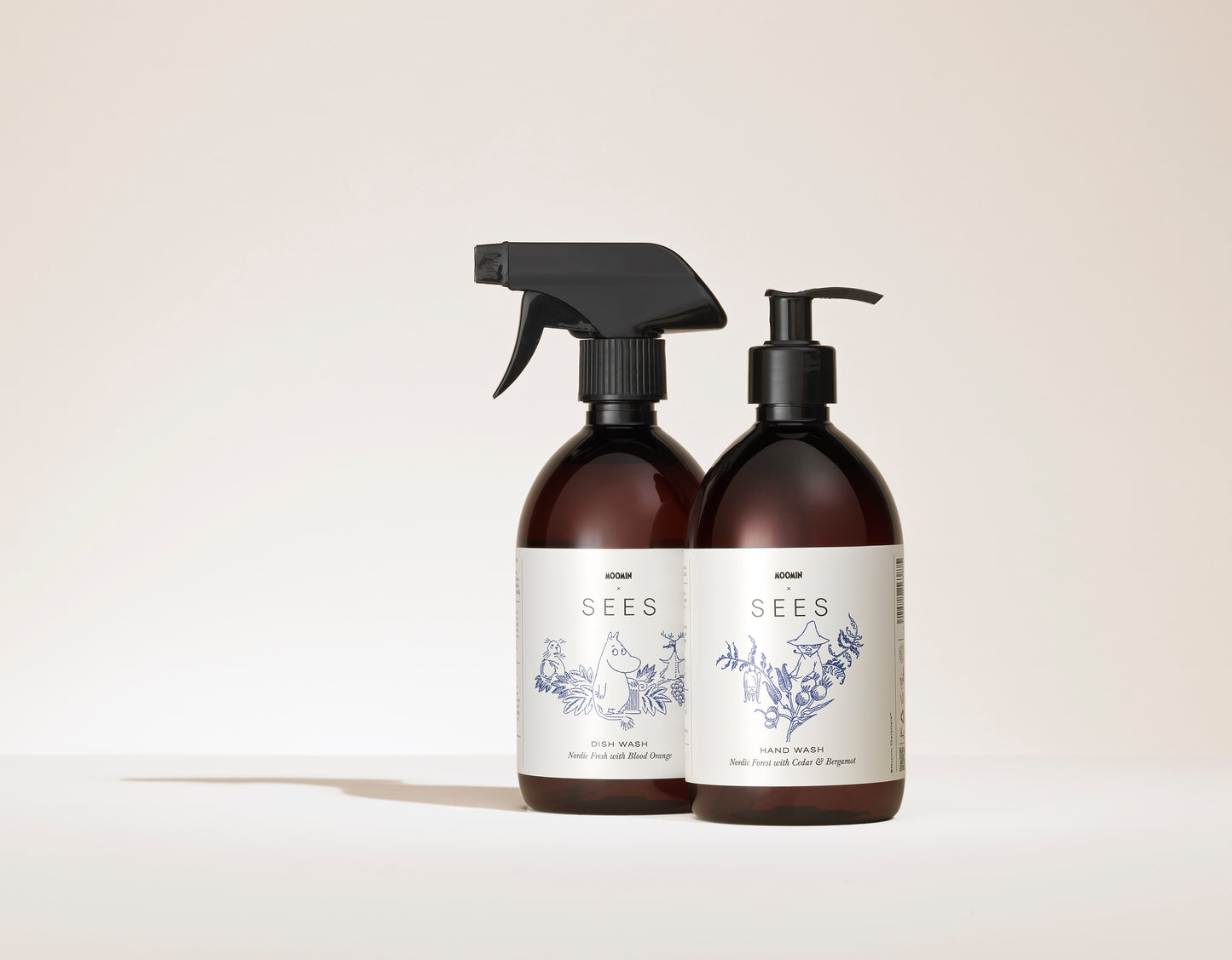 SEES x Moomin Hand Wash; kaksi ruskeaa pulloa valkoisin etikettein; toisessa suihkesuutin merkitty Dish Wash, toisessa pumppu merkitty Hand Wash; molemmissa etiketeissä sinisiä viivapiirroksia kukista ja eläimistä minimalistiseen tyyliin; pullot lieriömäisiä mattapinnalla; serif-typografiaa etikettitekstissä; tausta yksinkertainen ja neutraali; ei ihmisiä tai käsiä kuvassa.
