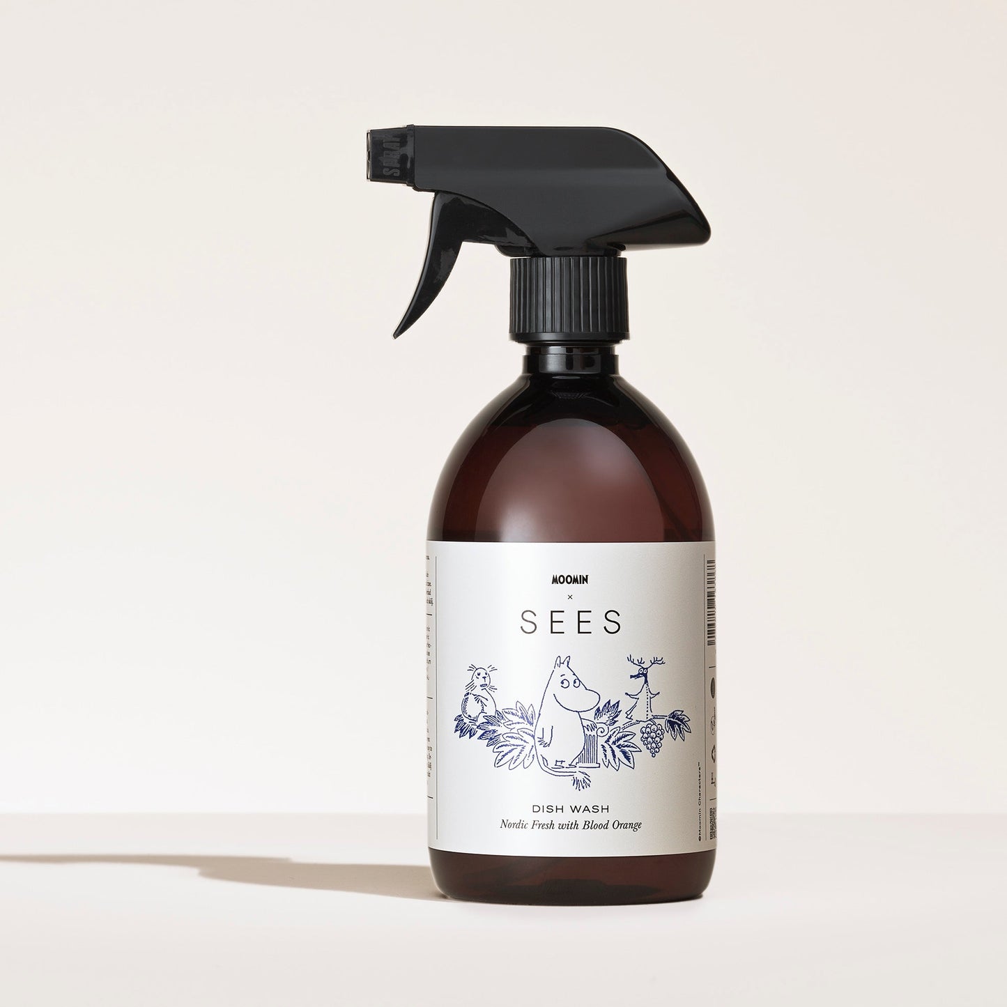 SEES x Moomin Dish Wash Spray; ruskea muovinen suihkepullo tekstillä SEES Dish Wash seisoo pystyssä mattapintaisella, vaaleanharmaalla neutraalilla pinnalla; valkoisessa kiiltäväpintaisessa etiketissä värikkäitä piirroshahmoja ja kasvikuvituksia; tuoksu Needle Peach and Blood Orange luettavissa; etiketin fonttityyli groteskia; ei käsiä tai ihmisiä kuvassa.
