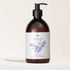 SEES x Moomin Hand Wash; ruskea muovinen pumppupullo mattapinnalla ja pyöristetyllä muodolla; etiketissä lukee SEES Hand Wash, näyttää yksinkertaisia viivapiirroksia kasveista ja muumihahmokuvituksen; typografia selkeää groteskia; lisätekstissä tuoksu: neulametsä setripuulla ja bergamotilla; tausta yksinkertainen ilman näkyviä käsiä tai rekvisiittaa.