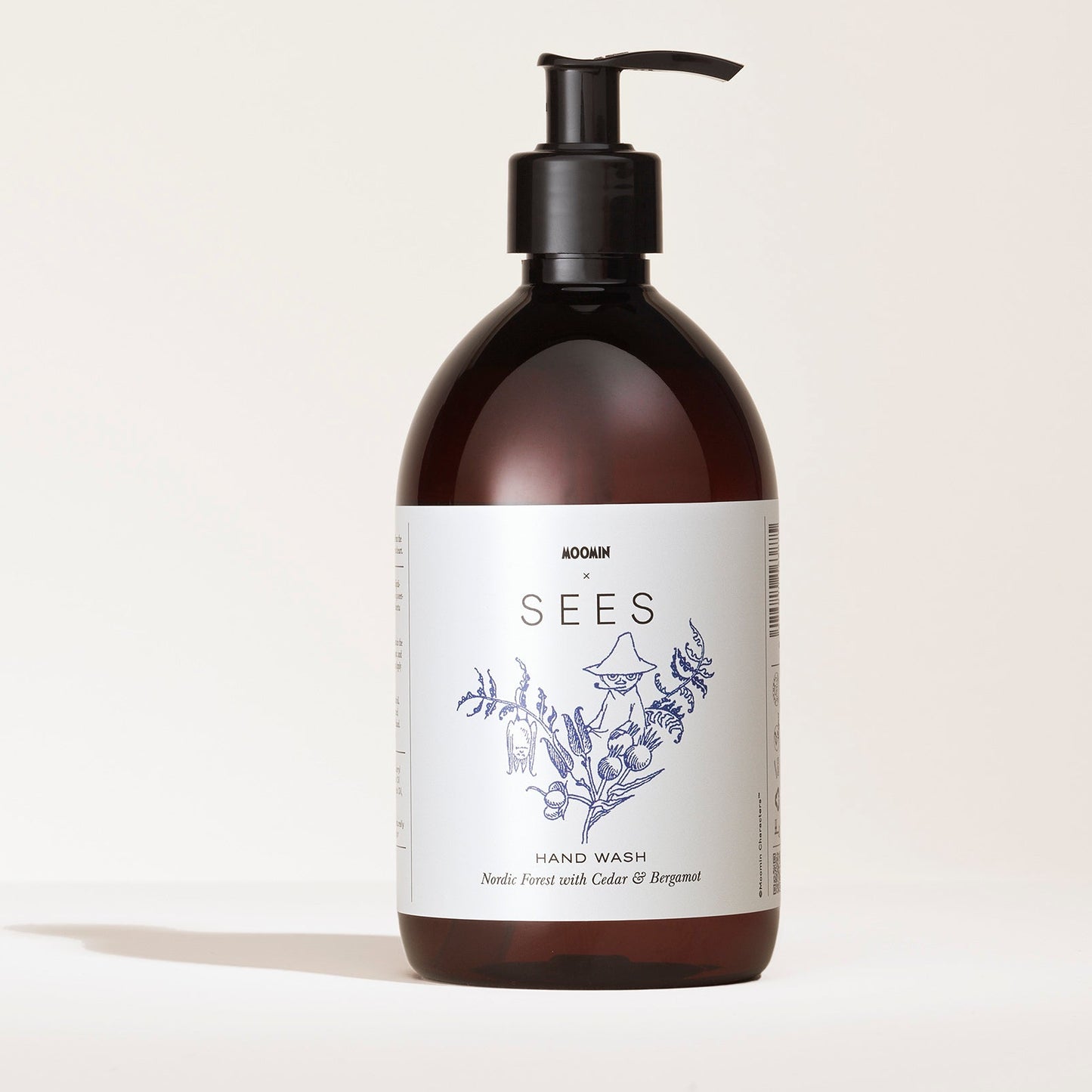 SEES x Moomin Hand Wash; ruskea muovinen pumppupullo mattapinnalla ja pyöristetyllä muodolla; etiketissä lukee SEES Hand Wash, näyttää yksinkertaisia viivapiirroksia kasveista ja muumihahmokuvituksen; typografia selkeää groteskia; lisätekstissä tuoksu: neulametsä setripuulla ja bergamotilla; tausta yksinkertainen ilman näkyviä käsiä tai rekvisiittaa.