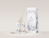SEES x Moomin Room Diffuser Nordic Summer; kirkas lasinen tuoksudiffuusori kolmella vaalealla puutikulla pystyssä; aseteltuna mattavalkoisen suorakaiteen muotoisen pakkauksen viereen, jossa sinisiä kasvikuvituksia ja mustaa groteskitekstiä SEES Scent Diffuser Nordic Summer; molemmat esineet vaalealla, yksinkertaisella taustalla; ei ihmisiä tai käsiä kuvassa.