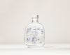 SEES x Moomin Room Diffuser Nordic Summer; kirkas lasipullo korkkikannella, tekstillä SEES SCENT DIFFUSER Nordic Summer, sinisiä kasvikuvituksia ja pieni piirroshahmo etuosassa; etiketin typografia sinistä groteskia; pinta kiiltäväpintainen; pullo lieriömäinen ja läpinäkyvä; ei ulkopakkausta näkyvissä; vaalea murrettu valkoinen tausta ilman lisärekvisiittaa tai ihmisiä; suoraviivainen tuotekuva silmän korkeudelta.