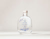 SEES x Moomin Room Diffuser Nordic Winter; kirkas lasipullo lieriömäisellä muodolla seisoo pystyssä yksinkertaista vaaleaa taustaa vasten; pullo suljettu luonnollisella korkkitulpalla; etiketissä teksti SEES Scent Diffuser siistillä groteskifontilla sekä sinisiä kasvikuvituksia; ei pakkausta tai muita esineitä näkyvissä; ei ihmisiä tai käsiä kuvassa.