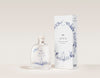 SEES x Moomin Room Diffuser Nordic Winter; kirkas lasinen tuoksudiffuusoripullo puisella korkilla ja leikkisin sinisin kuvituksin, aseteltuna vastaavan valkoisen pakkauksen viereen, jossa samankaltaista sinistä viivataidetta ja brändinimi SEES; molemmilla esineillä mattapinta; pullo lieriömäinen, pakkaus suorakaiteen muotoinen; laatikon typografia yksinkertainen ja groteskia; setti kuvattu pehmeää, neutraalia taustaa vasten ilman ihmisiä tai käsiä.