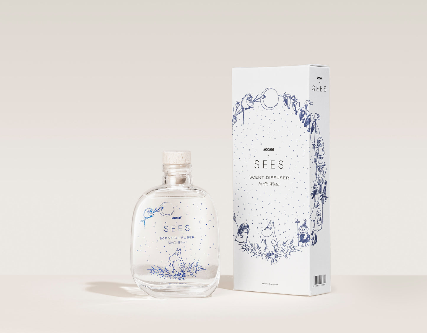 SEES x Moomin Room Diffuser Nordic Winter; kirkas lasinen tuoksudiffuusoripullo puisella korkilla ja leikkisin sinisin kuvituksin, aseteltuna vastaavan valkoisen pakkauksen viereen, jossa samankaltaista sinistä viivataidetta ja brändinimi SEES; molemmilla esineillä mattapinta; pullo lieriömäinen, pakkaus suorakaiteen muotoinen; laatikon typografia yksinkertainen ja groteskia; setti kuvattu pehmeää, neutraalia taustaa vasten ilman ihmisiä tai käsiä.