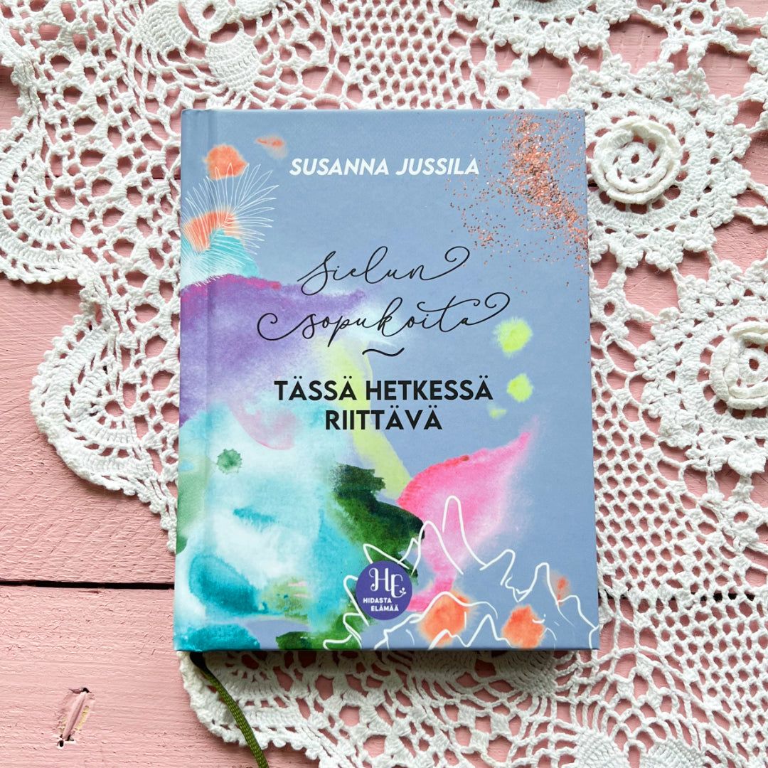 A colorful book titled Sielun Sopu Kohta Tässä Hetkessä Riittävä by Susanna Jussila rests on a pink wooden surface, with a delicate crocheted doily partially visible underneath. The book has a matte cover finish and displays bold, multicolor typography on the front. The shot is overhead with no people or hands shown.
