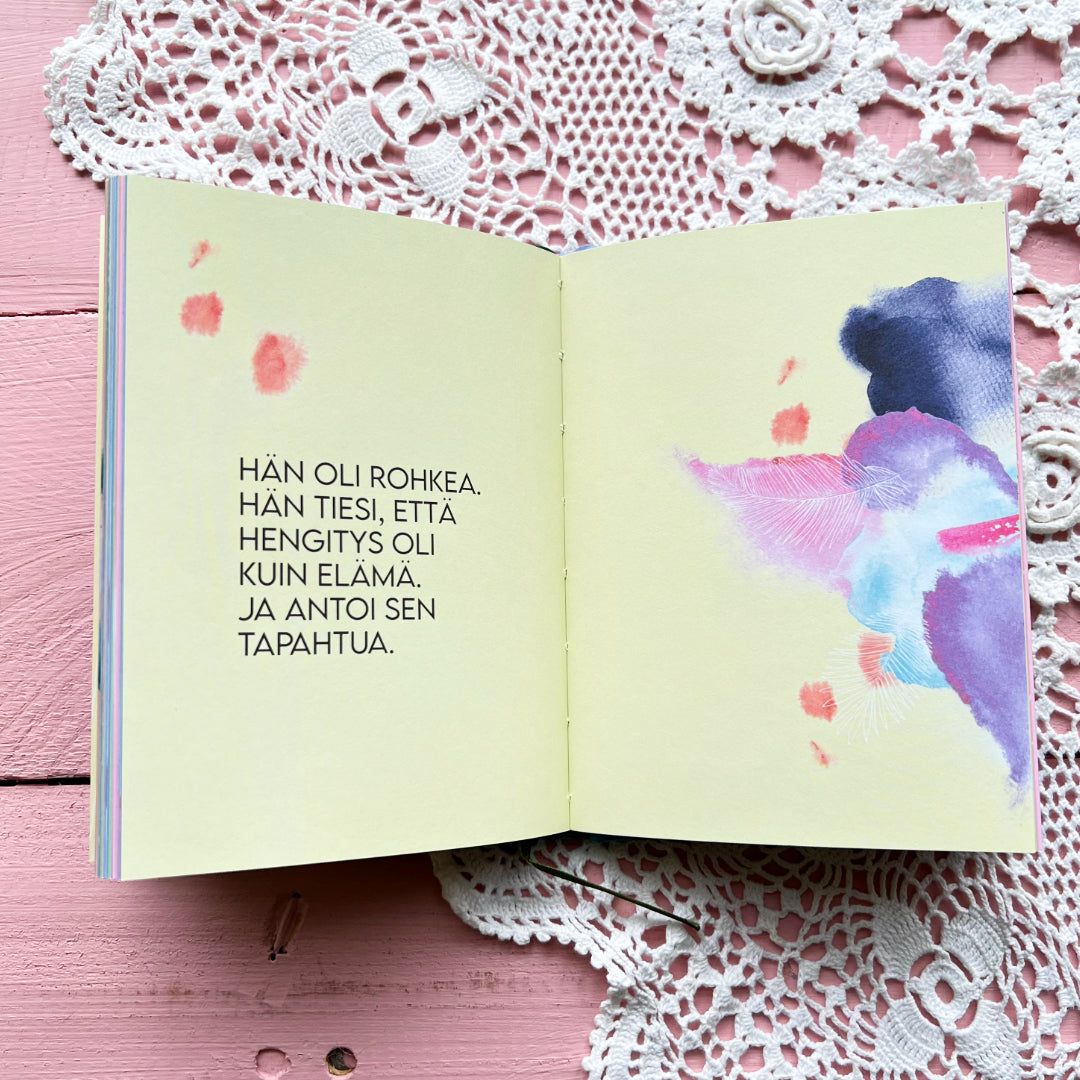 An open book rests on a pink wooden table with a white lace doily. The book has white pages; the left page contains printed Finnish text in a standard serif typeface, and the right page shows abstract watercolor art using soft purple, pink, and blue hues. No packaging is present. No people or hands are visible in the image. Overhead shot.