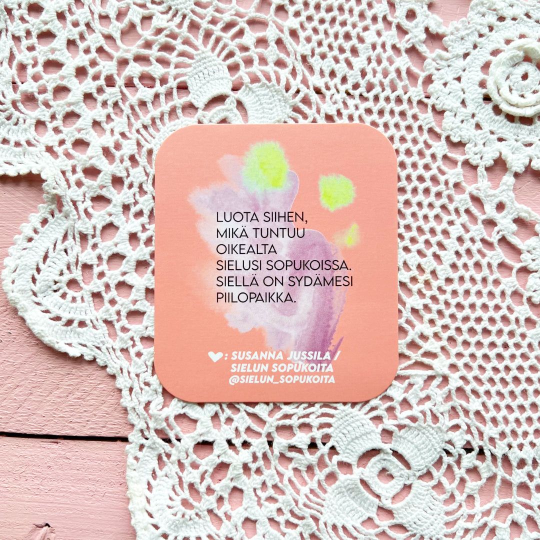 A pink coaster with Finnish text rests on a lacy white tablecloth and a pink wooden surface. The coaster is round, appears to be made of a rigid material with a matte finish, and displays soft pastel-colored artwork. The visible Finnish text uses clear sans serif typography. No people, hands, or packaging are present in the shot.