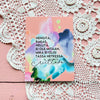 A pink card with Finnish text, vibrant watercolor designs, and elegant cursive writing rests on a pink wooden surface with white crocheted doilies. The card is rectangular, made of matte cardstock, and features multicolored watercolor elements. Typography combines printed Finnish text and decorative cursive. No packaging or people are present in the shot.