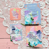 Books and cards with pastel watercolor designs and Finnish text are arranged on a pink wooden surface with white lace doilies. Covers have matte finishes; most items are rectangular. Typography is mostly sans serif, in medium to large size. Legible words include motivational and caring messages in Finnish. No people or hands are visible; flat lay shot.