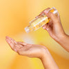 A person pours a hydrating skincare serum from a transparent bottle into their open palm, against a soft yellow background. The bottle is clear plastic or glass with a cylindrical shape and glossy finish; any label text is not legible. The person’s hand holds the bottle upside down above the open palm. This is a close-up shot with no other objects or setting visible.