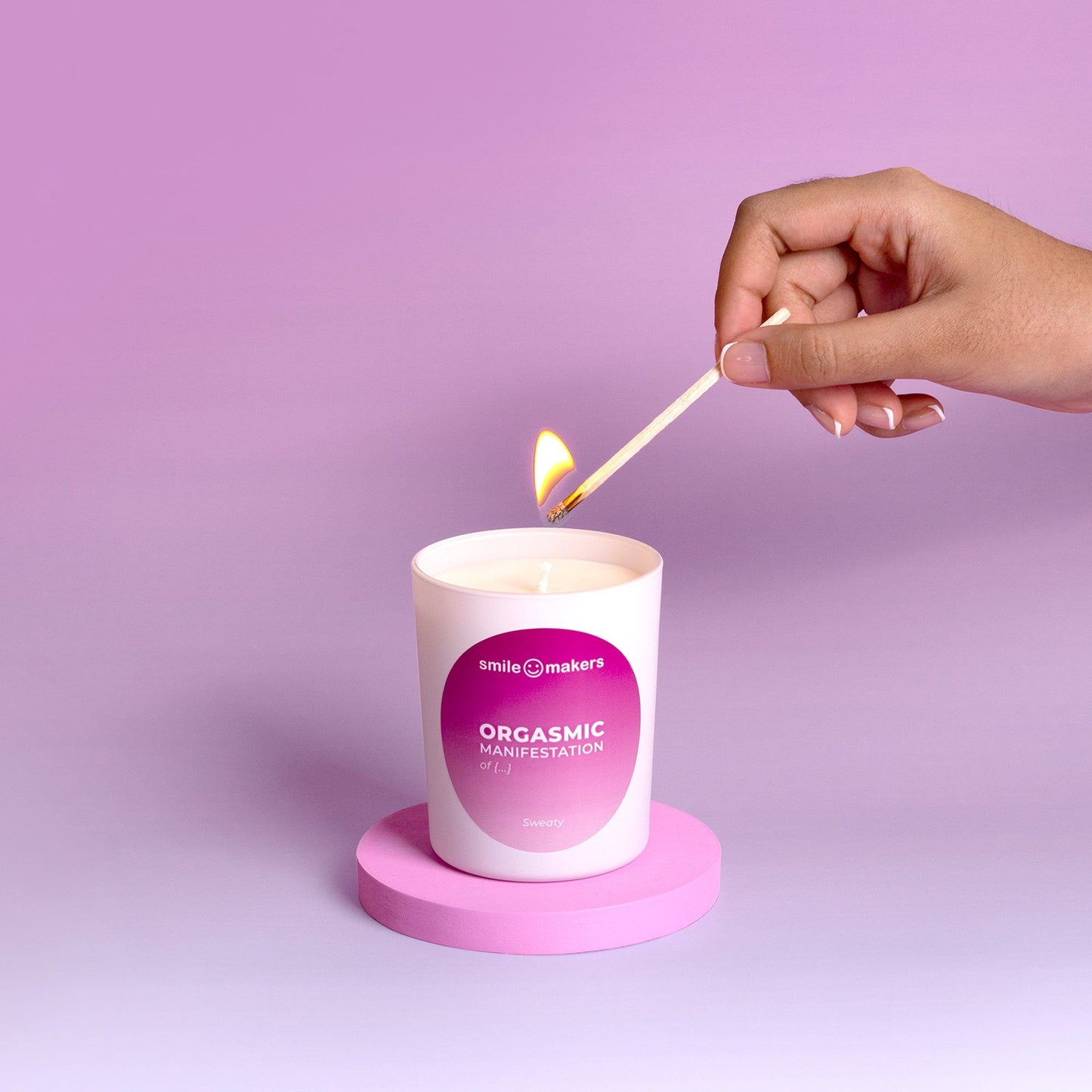 A hand lights a white candle labeled Orgasmic Manifestation, set on a pink stand against a dreamy pink background. The cylindrical candle has a matte finish and sits upright. The product label displays clear, sans serif text in black. Background is uniformly soft pink with smooth gradient effect. Close-up shot, only one hand visible engaging with the candle.