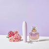 A white vibrator, a pink perfume bottle, a diamond ring, and pink flowers are artfully arranged on a white surface with a purple background. The vibrator is smooth with rounded edges and no visible branding. The perfume bottle is cube-shaped glass with a matte silver cap. The ring has a large central clear stone in a metal band. Flowers have full pink petals and green stems. No people or hands are present. No legible label text is visible. Shot is overhead top-down view with soft shadows.