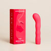 Smile Makers The Romantic G-pistevibraattori; vaaleanpunainen kaareva vibraattori pystyssä punaisen laatikon vieressä, jossa abstrakti kasvokuvitus ja teksti "The Romantic Sensuous Vibrator by Smile Makers"; vibraattori sileän mattapintainen pyöristetyllä kärjellä; laatikossa yksinkertaista viivakuvitusta ja mustaa groteskitekstiä punaisella pohjalla; ei ihmisiä tai käsiä, neutraali tausta.