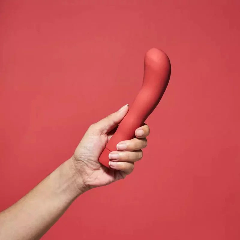 Smile Makers The Romantic G-pistevibraattori; käsi pitelee punaista kaarevaa esinettä, jonka pinta on kiiltävä ja sileä ilman näkyviä merkintöjä; muoto pitkulainen kapenevin päin; ei pakkausta; tausta yksivärisen punainen; kuvassa vain yksi käsi, joka pitää esinettä otteessaan alta.