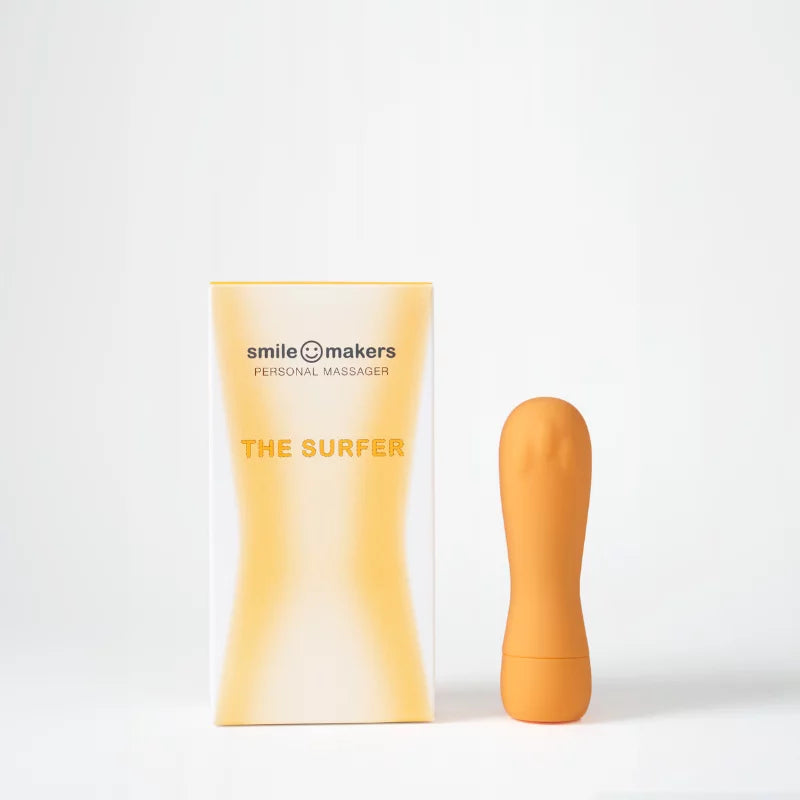 Smile Makers The Surfer Klitorisvibraattori; keltainen pahvilaatikko tekstillä "Smile Makers Personal Massager The Surfer" sans-serif-fontilla pystyssä; vieressä oranssi silikonivibraattori pyöristetyllä muodolla ja mattapinnalla pystyasennossa; molemmat saumattomalla valkoisella taustalla; ei ihmisiä tai käsiä kuvassa.