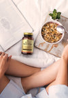Solgar Female Multivitamin: henkilö istuu sängyllä avoimen kirjan, granolakulhon lusikan kanssa, pienen ruukkukasvin ja Female Multivitamin -pullon vieressä; monivitamiinipullo on sylinterimäinen, valkoista mattamuovia, kirkkaalla etiketillä tekstillä "Female Multivitamin" suurella groteskikirjasimella; kuva on otettu ylhäältä luonnonvalossa neutraaleilla vuodevaatteilla.