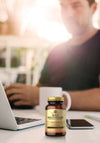 Solgar Male Multivitamin: muovipullo miesten monivitamiinikapseleita mattaetiketillä seisoo pystyssä puisella työpöydällä; etiketissä selkeä groteskiteksti "Men's Multivitamin" sinisin ja valkoisin värein; lähellä musta älypuhelin ja hopeanvärinen kannettava tietokone; taustalla epätarkkana henkilö pitelee valkoista mukia työskennellen avoimen kannettavan äärellä.