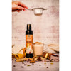 A hand sprinkles powder through a fine metal sieve over a clear glass mug filled with brown chai. Next to the mug is an amber glass bottle with a white label reading “Chai Bio-Sirup” in bold black sans-serif font. Whole spices, a stainless steel measuring cup, and a metal spoon rest on a light-colored tabletop. The shot is set indoors with even lighting. A hand sprinkles powder through a sieve over a glass mug of chai beside a bottle labeled Chai Bio-Sirup. Fragrant spices, a measuring cup, and a spoon are