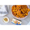 Pyöreä valkoinen keraaminen kulho sisältää kultaisenruskeita perunalastuja, joissa näkyy maustetta. Vieressä on pieni valkoinen kulho kermaista kastiketta silputuin yrttein ja maustein. Suorakulmainen laatikko, jossa lukee Chakali Bhakar lihavoidulla tummalla tekstillä, seisoo niiden takana. Asetelma lepää mattaharmaalla pinnalla raidallisen liinan peitossa; ihmisiä ei näy.