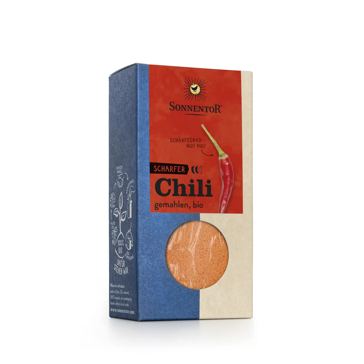 Laatikko Sonnentor Scharfer Chili, gemahlen, bio -tuotetta, jonka etupuolella on eloisa punaisen chilipippurin kuva ja pyöreä ikkuna, joka näyttää luomuchilijauheen sisällä. Suorakulmainen pahvilaatikko on viimeistelty mattana rohkein punaisin, sinisin ja beigein värein. Etiketissä on mustaa pääteviivatonta tekstiä. Ihmisiä tai käsiä ei näy; tausta on valkoinen, kyseessä on suoraviivainen tuotekuva.