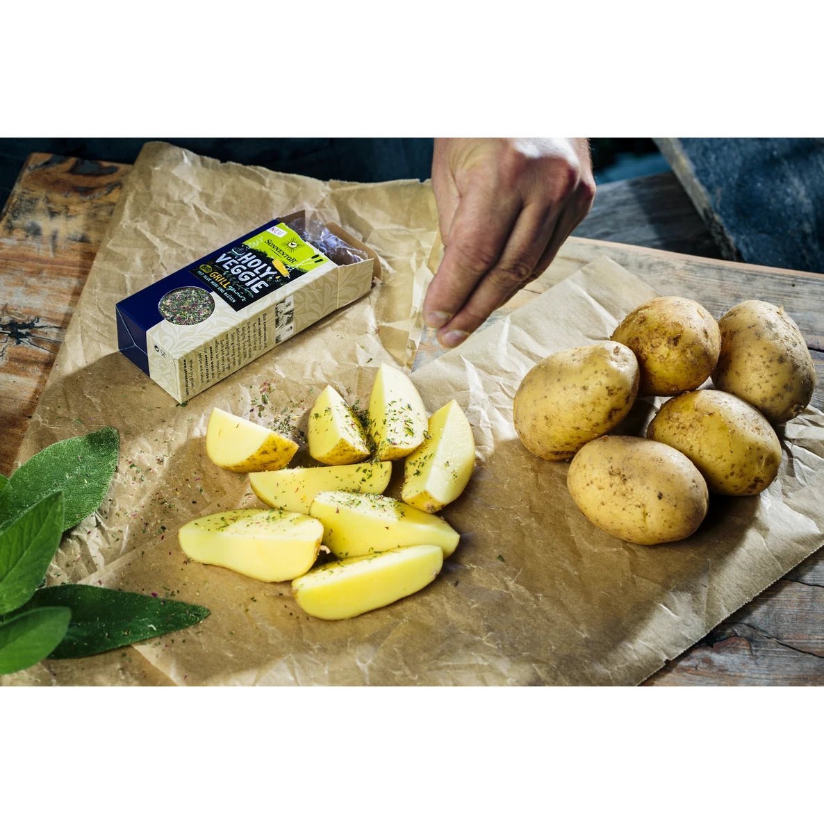 Käsi ripottelee yrttejä viipaloitujen perunoiden päälle ruskealla paperilla, kuorimattomien kokonaisten perunoiden, salvianlehtien ja suorakulmaisen laatikon vieressä, jossa lukee flavorful veggie stock lihavoidulla pääteviivattomalla fontilla. Esineet on aseteltu mattapintaiselle puupinnalle näkyvin syin. Ylhäältä otettu kuva, joka rajaa kaikki esineet tiiviisti yhteen luonnonvalossa.