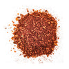 Kasa hienoksi jauhettua punaruskeaa paprikaa pienine epätasaisine paloineen on siroteltu valkoiselle taustalle. Paprika on löyhää ja pakkaamatonta, mattapintaista, ilman näkyvää pakkausta tai etikettejä. Tausta on pelkistetty ja valkoinen, ja kuva tarjoaa suoran näkymän ylhäältä. Ihmisiä tai typografiaa ei näy.
