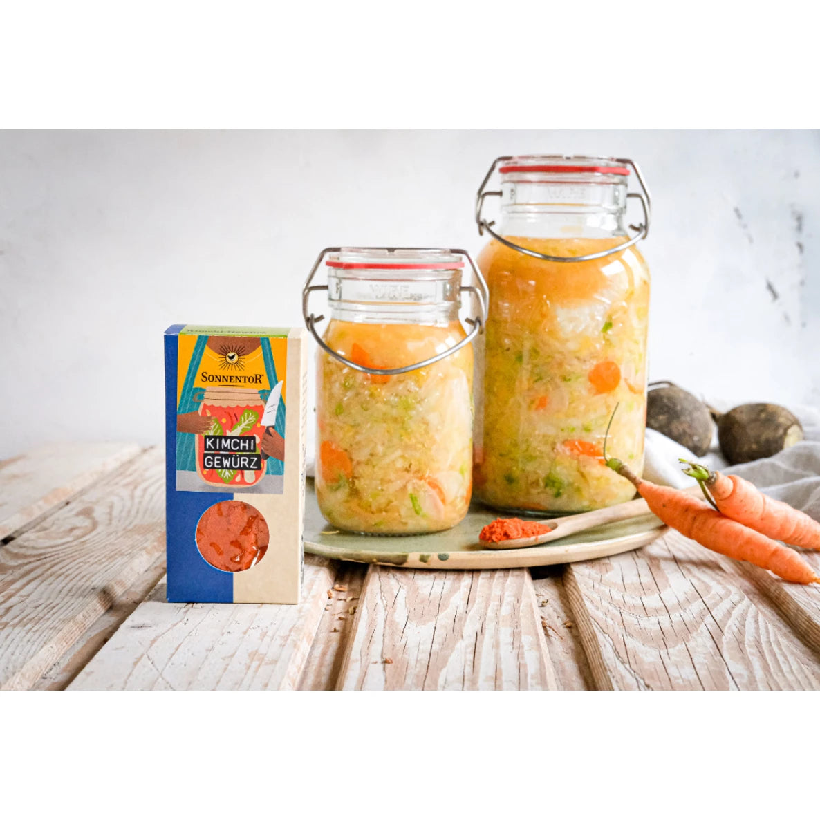 Kaksi purkkia kotitekoista kimchiä näkyvine porkkana- ja vihannespaloineen on aseteltu suorakulmaiselle tarjottimelle painetun pahvilaatikon vieressä, jossa lukee kimchi spice mix selkeällä pääteviivattomalla tekstillä. Kohtauksessa on kokonaisia irtoporkkanoita, metallinen lusikka täynnä punaista mausteenjauhetta, sekalaisia vihanneksia, ja se lepää käsittelemättömällä puisella pöytäpinnalla. Kuva on otettu ylhäältä.