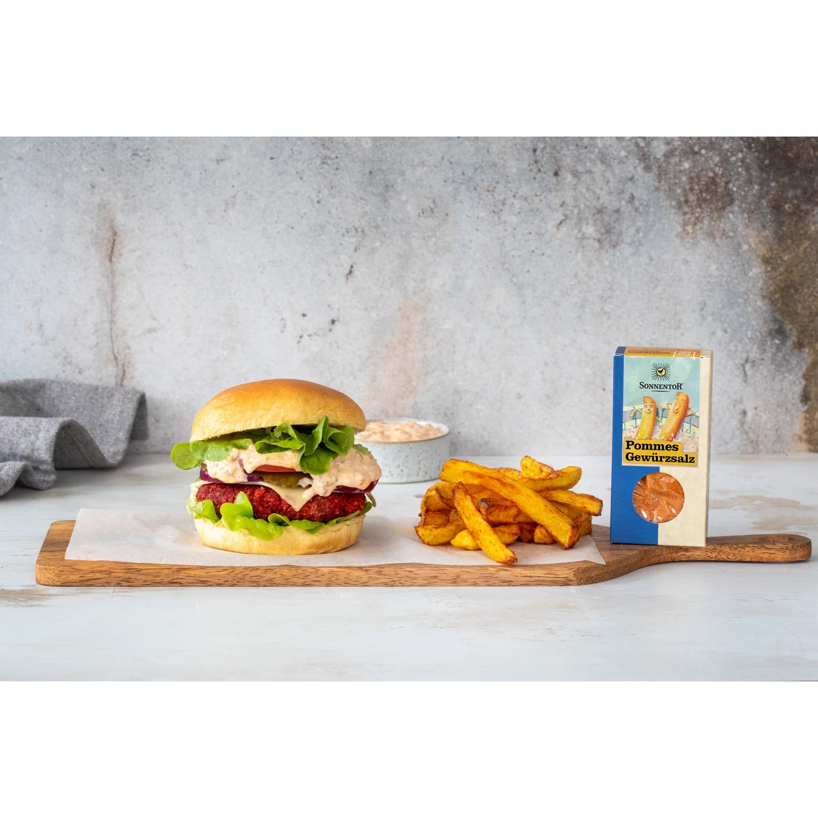 Hampurilainen salaatilla, tomaatilla ja kastikkeella seisoo suorakulmaisella puulaudalla ranskalaisten kasan vieressä. Laatikko, jossa lukee Fries Seasoning lihavoidulla pääteviivattomalla fontilla, on ranskalaisten vieressä. Taustalla näkyy harmaa kangasliina ja pieni valkoinen kulho. Ihmisiä tai käsiä ei näy. Ylhäältä otettu kuva neutraalissa valaistuksessa.