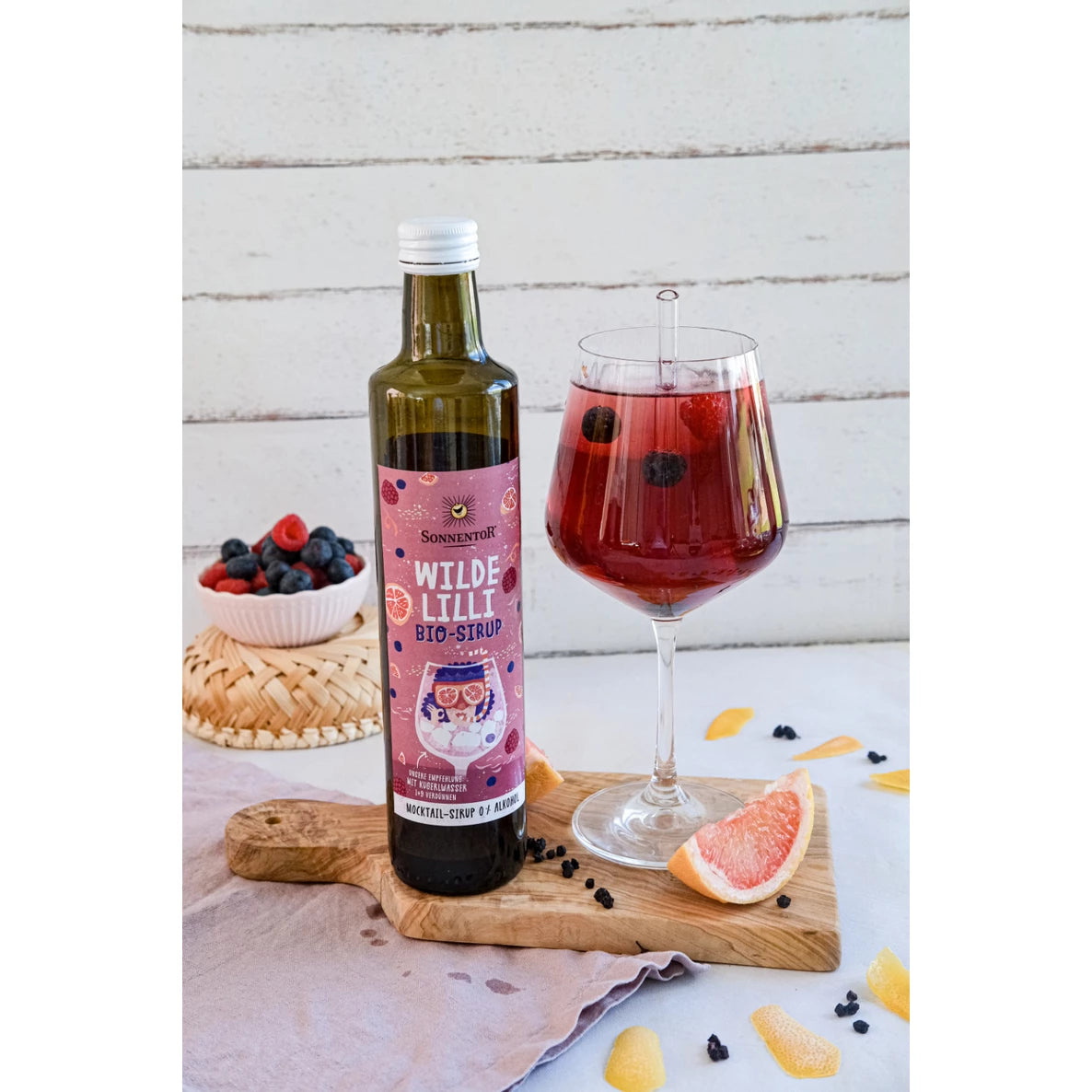 Pullo Sonnentor Wilde Lilli Bio-Sirup -siirappia, jossa on tummaa nestettä ja vihreä korkki, seisoo suorakulmaisella puulaudalla. Lasipullossa on mattapintainen paperietiketti, jossa lukee selkeästi brändi ja tuotenimi lihavoidulla, yksinkertaisella fontilla. Vieressä on kirkas lasi punaviolettia marjajuomaa jääpaloin ja marjoin. Valkoinen kulho sisältää sekalaisia marjoja; lähellä on irtomarjoja ja puolikas greippiviipale vaalealla pinnalla ilman koristeita tai kuvioita. Ihmisiä tai käsiä ei näy.