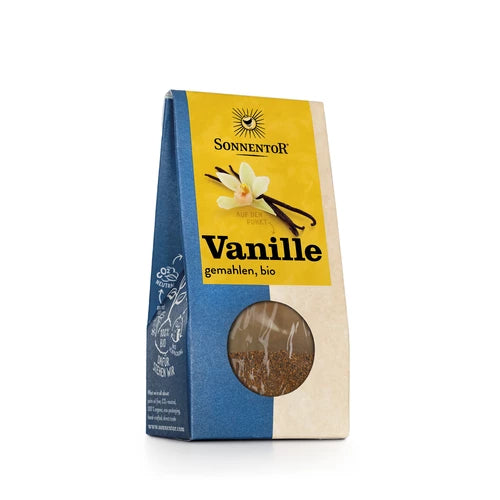 A blue and yellow Sonnentor package labeled Vanille gemahlen, bio features an image of vanilla flowers and pods, along with a small window displaying the ground vanilla inside. Rectangular cardboard box with matte finish; main colors are cobalt blue on the left, pale yellow on the right, and dark blue at top. Package front shows printed illustration of three yellow vanilla flowers and green leaves beside two brown pods. A round transparent plastic window near bottom right reveals dark brown powder inside.