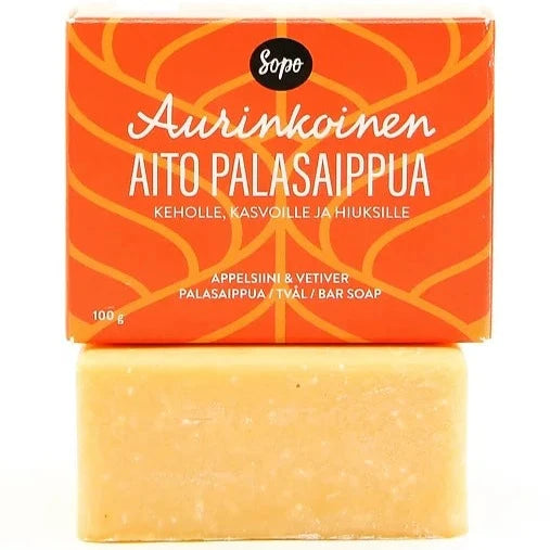 Sopo Aurinkoinen Aito Palasaippua; oranssi saippuapala mattapinnalla lepää suorakaiteen muotoisen laatikon vieressä; laatikossa teksti "Aurinkoinen Aito Palasaippua" lihavoiduin groteskisuuraakkosin; pakkaus pääosin valkoinen ja oranssi, pienempi suomenkielinen teksti kuvaa tuoksun appelsiiniksi ja vetiveriksi; molemmat tuotteet yksinkertaisella, neutraalilla taustalla tasaisessa valaistuksessa; ei ihmisiä eikä käsiä näkyvissä.