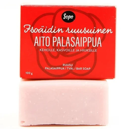 Sopo Isoäidin Ruusuinen Aito Palasaippua; vaaleanpunainen saippuapala punaisen pakkauksensa edessä, jossa suomenkielinen teksti "Aito Palasaippua" ja "100 g"; saippua suorakulmainen hieman pyöristetyin reunoin ja mattapinnalla; punaisessa laatikossa valkoista groteskitekstiä etusivulla; tausta neutraali ja siisti, ei ihmisiä eikä käsiä näkyvissä, kuva suoraan edestä.