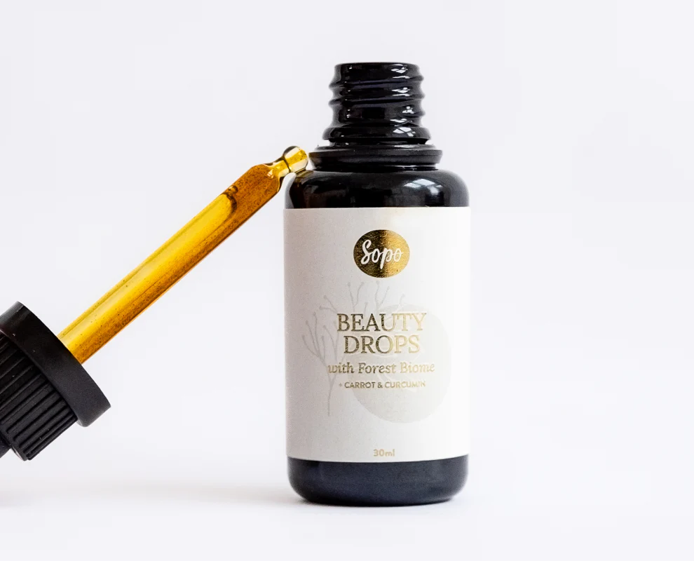 Sopo Kauneusöljy; tumma lasipullo tekstillä "Sopo Beauty Drops with Forest Biome" valkoisella taustalla, vieressä tippa täynnä kultaisenkeltaista nestettä; pullo sylinterimäinen kiiltäväpintainen ja musta kierrekorkki; luettavassa etiketissä yksinkertaista groteskityyppiä; ei ihmisiä eikä käsiä näkyvissä; kuva suoraan edestä tasaisessa valaistuksessa.