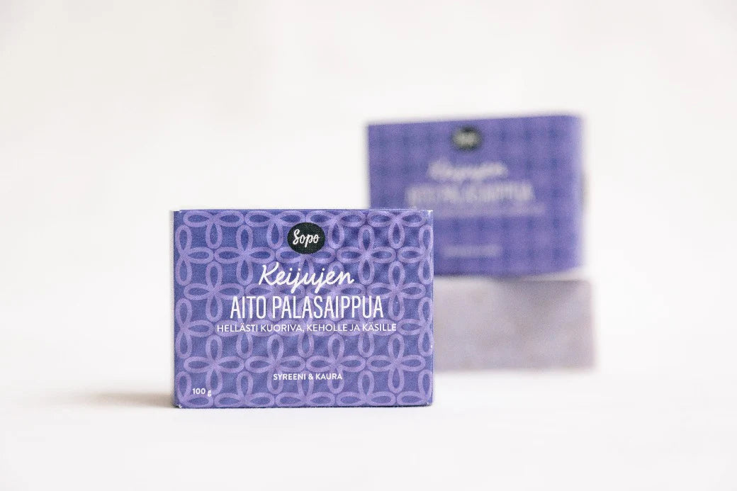 Sopo Keijujen Aito Palasaippua; violetti saippuapala mattapinnalla suorakaiteen muotoisen pahvilaatikon sisällä, jossa monivärinen geometrinen kuvio ja etiketti "Keijujen Aito Palasaippua" groteskitekstillä; sekä saippua että pakkaus suorakulmaisia; taustalla toinen kuvioitu saippualaatikko ja vastaava saippuapala pehmeästi epätarkkoina valkoisella pinnalla.