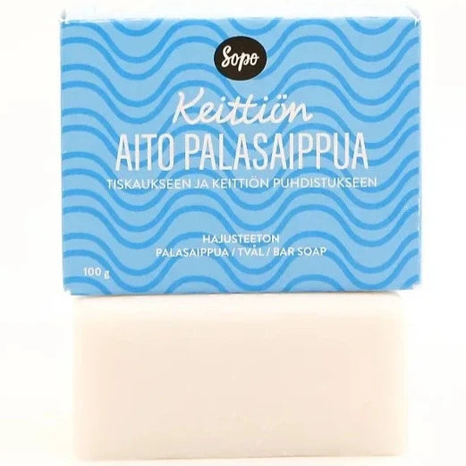 Sopo Keittiön Aito Palasaippua Hajusteeton; sininen ja valkoinen pakkaus tekstillä "Keittiön Aito Palasaippua" sijaitsee pakkauksesta poistetun valkoisen saippuapalan yläpuolella; suorakaiteen muotoisessa laatikossa aaltoilevia sinisiä viivoja mattapinnalla, kaikki teksti lihavoitua suomenkielistä groteskia; saippua alla suorakulmainen tasaisin sivuin ja sileine reunoineen; molemmat tuotteet yksinkertaisella, heijastamattomalla pinnalla; ei ihmisiä eikä käsiä näkyvissä.