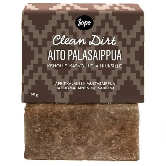 Sopo Metsäbiomi Palasaippua Clean Dirt; ruskea saippuapala mattapinnalla, osittain kääritty kuvioidulla paperikääreellä; kääreessä lohkomainen groteskiteksti "Sopo Clean Dirt Aito Palasaippua for body, face, and hair, made with African black soap and enriched with Finnish forest biome ingredients, 60g"; ei ihmisiä, käsiä eikä rekvisiittaa näkyvissä; tausta vaalea ja yksinkertainen, kuva läheltä.