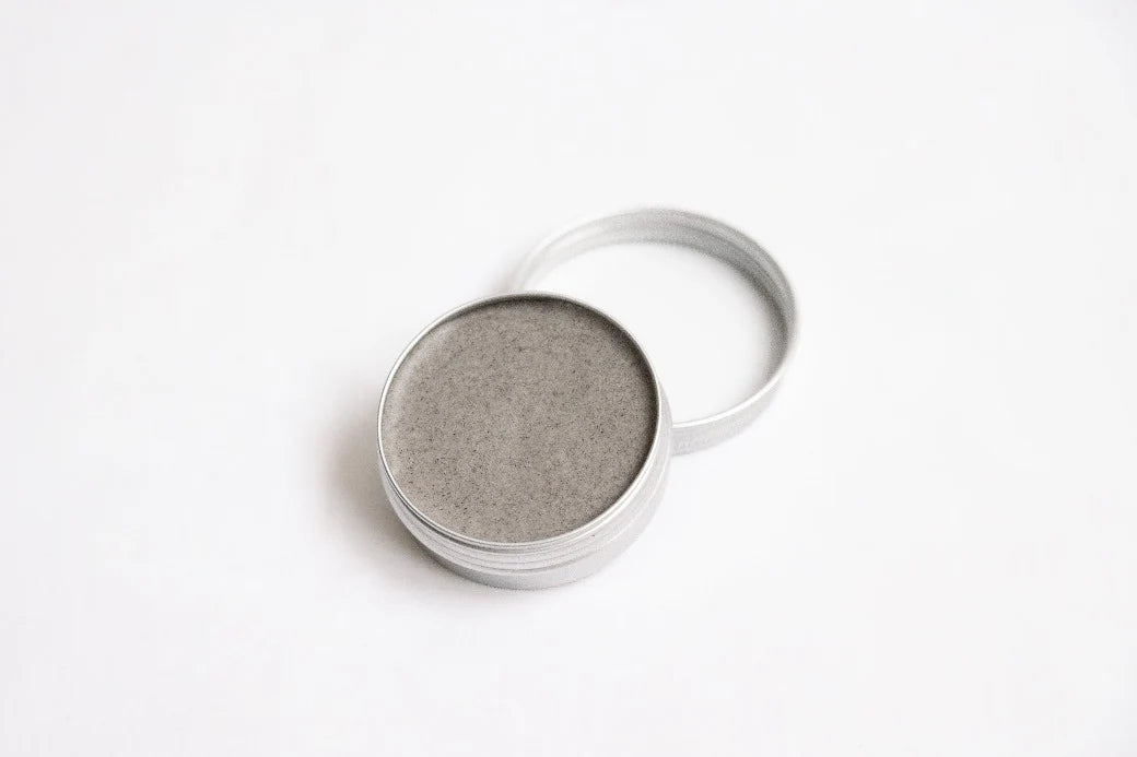 Sopo Mustikkainen Metsäbiomi; pieni pyöreä avoin metallitölkki, jossa sisällä harmaa kiinteä voide, seisoo valkoisella pinnalla; kansi aseteltu tölkin taakse; sekä tölkki että kansi mattahopeanvärisiä ja sileäpintaisia ilman näkyvää brändäystä tai tekstiä; ei ihmisiä eikä käsiä näkyvissä; asetelma minimalistinen tasaisella valaistuksella.