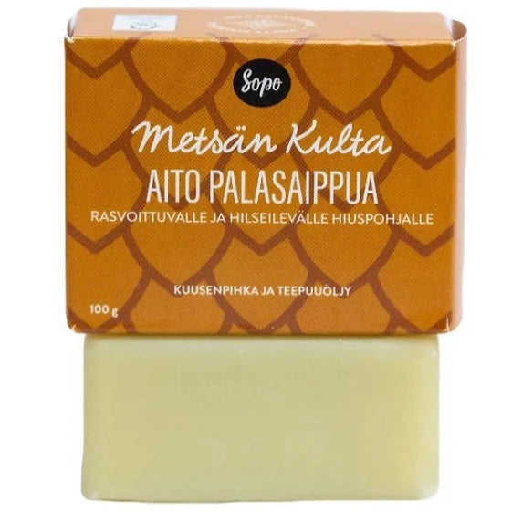 Sopo Metsän Kulta Aito Palasaippua; ruskea ja oranssi suorakaiteen muotoinen pahvilaatikko tekstillä "Metsän Kulta Aito Palasaippua" lepää vaaleankeltaisen suorakaiteen muotoisen kiinteän saippuapalan päällä; laatikossa yksinkertaista, groteskista suomenkielistä tekstiä kuvaten ainesosia ja käyttöä; molemmilla esineillä mattapinta; ei ihmisiä eikä käsiä näkyvissä, tausta neutraali.