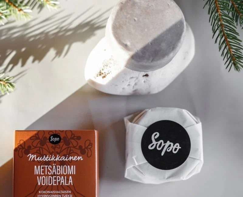 Sopo Mustikkainen Metsäbiomi; saippuapala, kääreeseen paketoitu Sopo-tuote ja ruskea laatikko suomenkielisellä tekstillä on aseteltu valkoiselle pinnalle; saippuapala suorakulmainen ja kermanvalkoinen; kääritty tuote taitellussa paperipakkauksessa yksinkertaisella painatuksella; ruskea laatikko mattapintaista pahvia mustalla groteskitekstillä; kuvan kulmissa näkyvissä mäntyoksia, luoden luonnollisen tai juhlallisen tunnelman.