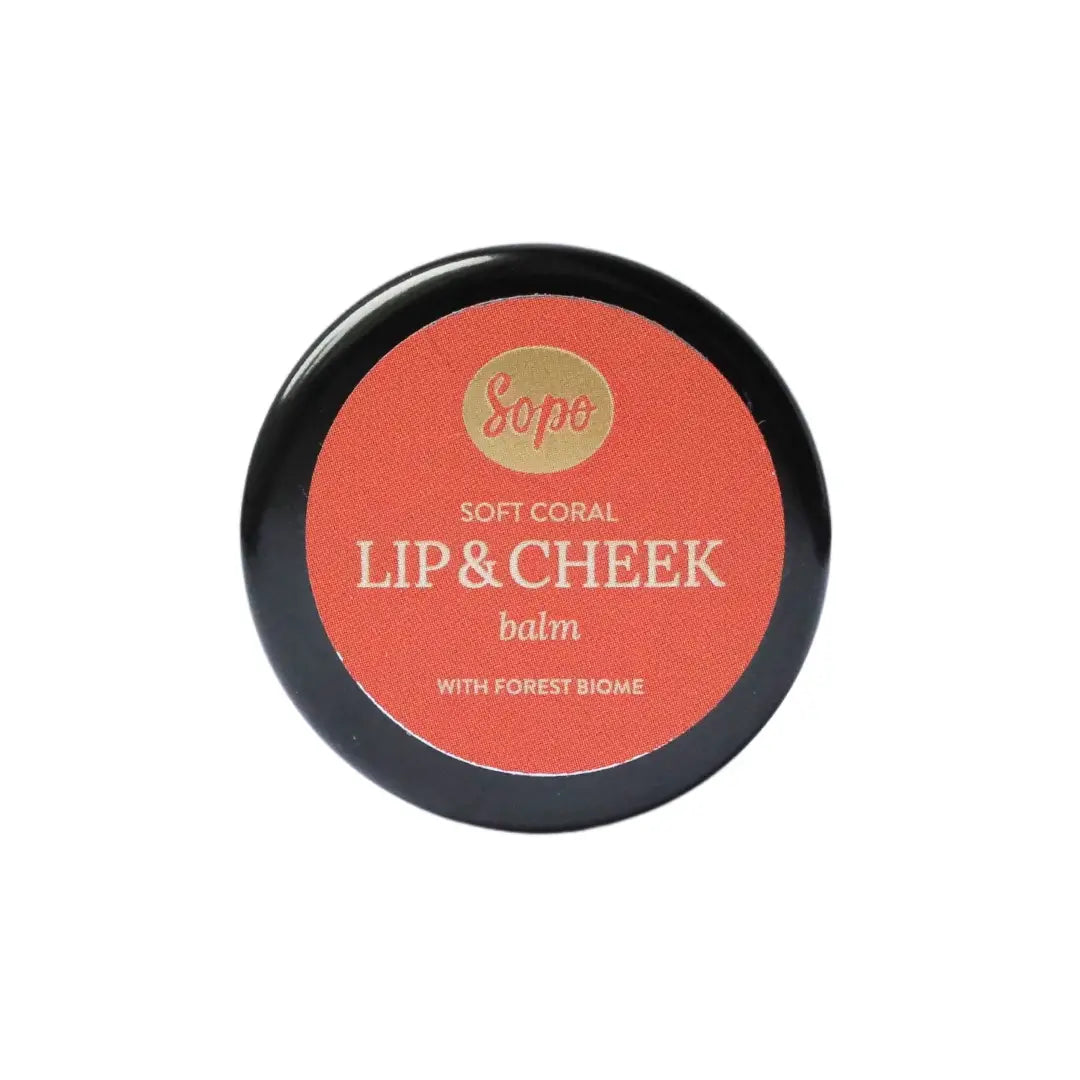 Sopo Sävyttävä Metsäbiomi Huulivoide & Poskipuna; pyöreä rasia tekstillä "Sopo Soft Coral Lip & Cheek Balm with Forest Biome", korallinvärinen kansi ja valkoista groteskitekstiä; rasian pohja mattapintainen ja luonnonvalkoinen; ei muita kuvioita tai koristeellisia yksityiskohtia näkyvissä; tausta valkoinen; kuvassa vain tuote, ei ihmisiä eikä käsiä näkyvissä.