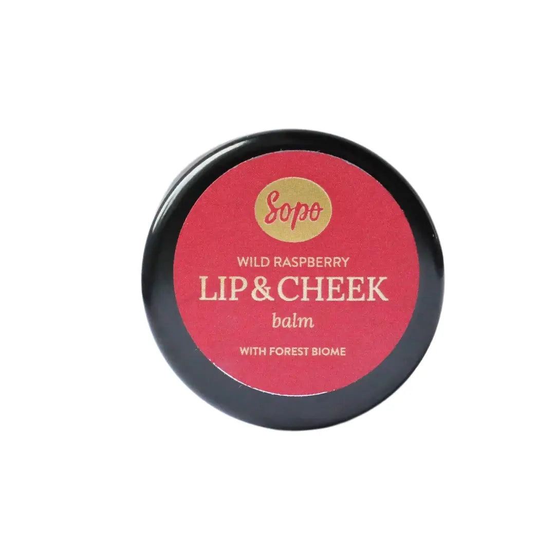 Sopo Sävyttävä Metsäbiomi Huulivoide & Poskipuna; pyöreä musta rasia punaisella etiketillä tekstillä "Sopo Wild Raspberry Lip & Cheek Balm", sisältää metsäbiomiuutteita; rasia läpikuultamaton mattapinnalla, kansi litteä; punainen paperietiketti peittää suurimman osan kannen pinnasta lihavoidulla valkoisella groteskitekstillä; tuote yksinkertaisella, vaaleasävyisellä alustalla; ei ihmisiä kuvassa.