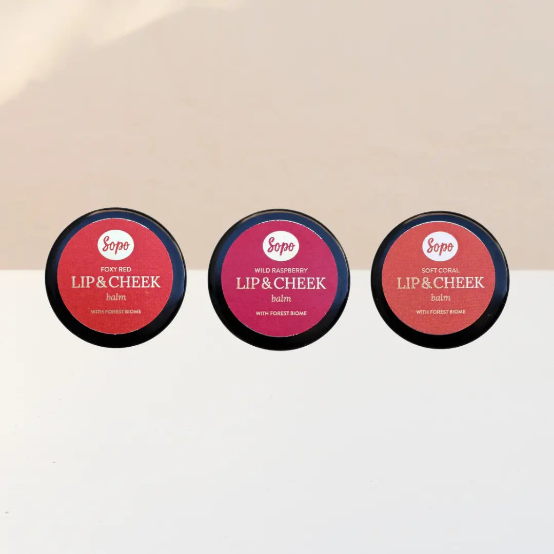 Sopo Sävyttävä Metsäbiomi Huulivoide & Poskipuna; kolme pyöreää Sopo Lip & Cheek Balm -rasiaa sävyissä Foxy Red, Wild Raspberry ja Soft Coral, aseteltuna riviin vaaleaa taustaa vasten; jokainen rasia metallinen ja mattapintainen, etiketöity selkeällä groteskitekstillä sekä brändi että sävy tunnistettavissa; ei ihmisiä eikä käsiä näkyvissä; suoraviivainen tuotekuva.