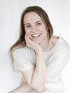 A woman with long brown hair smiles brightly, resting her chin on her hand. She wears a light-colored shirt. The background is plain white. The image shows only her upper body in a straightforward portrait shot, with no products or packaging visible. There are no labels, typography, or additional props present in the scene.